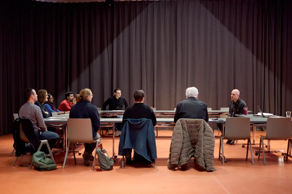 Menschen sitzen in einem Rechteck an Tischen. Der Mann am Tischkopf spricht zu den Menschen. Sie hören gespannt zu.