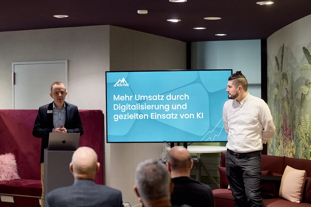 Zwei Männer halten vor einem Publikum eine Präsentation. Auf dem Bildschirm hinter ihnen befindet sich ihre Präsentation.