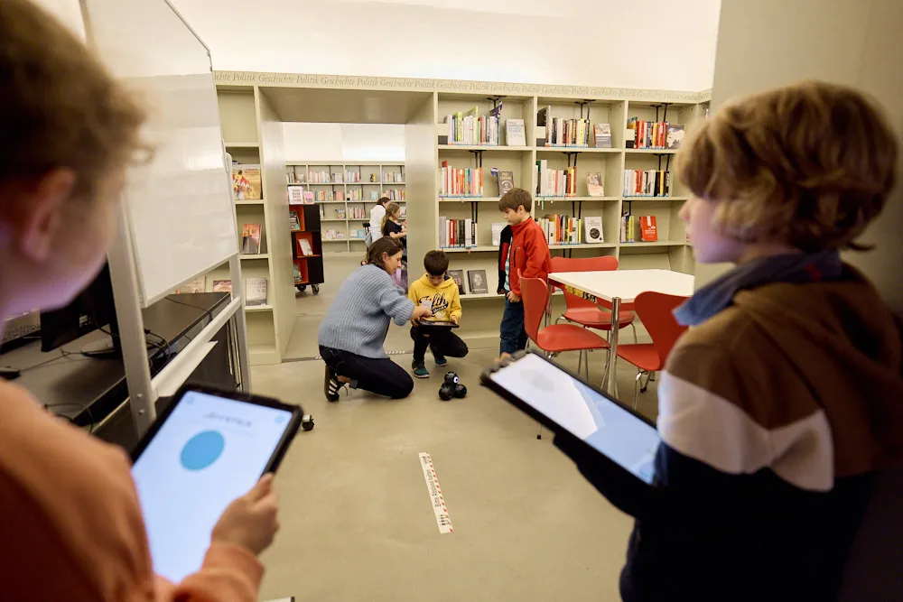 Kinder in einer Bibliothek bedienen mit Tablets kleine Roboter.