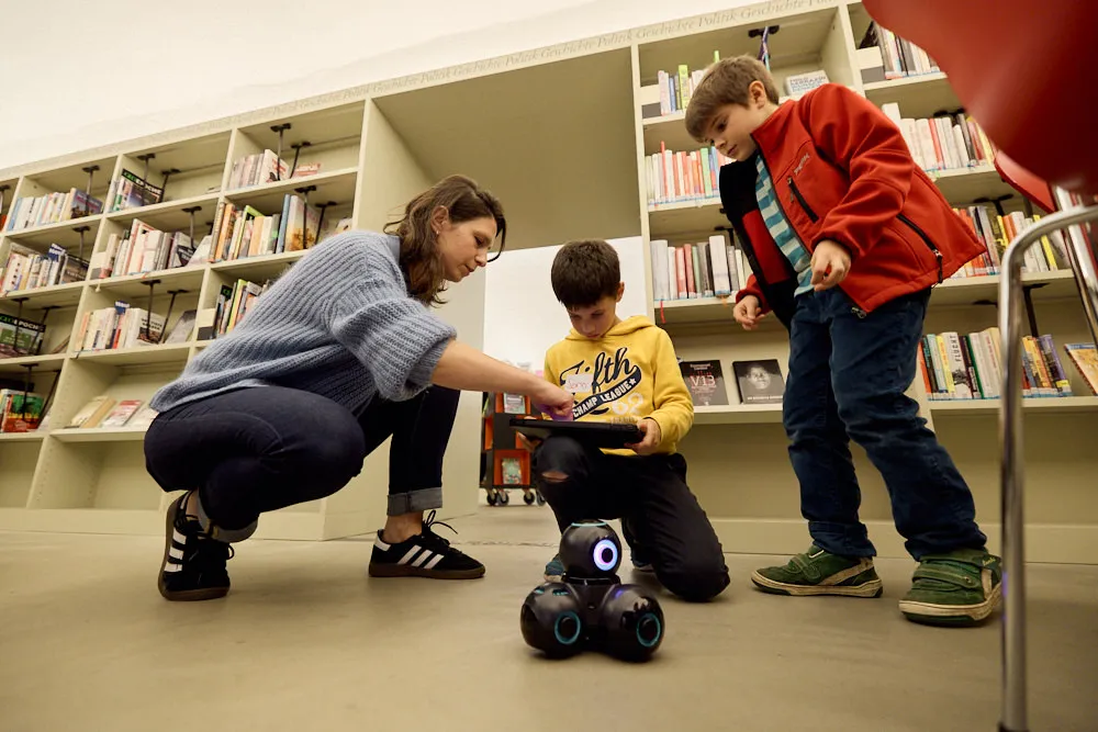 Eine Frau mit zwei Kindern in einer Bibliothek bedienen mit einem Tablet einen kleinen Roboter.