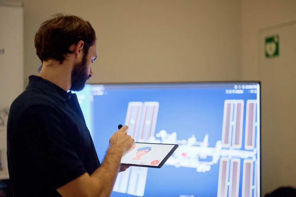 Ein Mann hält ein Tablet in der Hand. Vor ihm ist ein grosser Bildschirm.