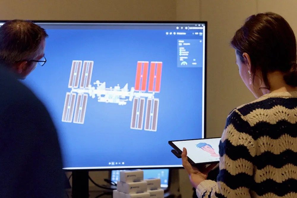 Eine Frau hält ein Tablet. Neben ihr steht ein Mann. Vor ihnen ist ein Bildschirm. Der Mann schaut auf dem Bildschirm.