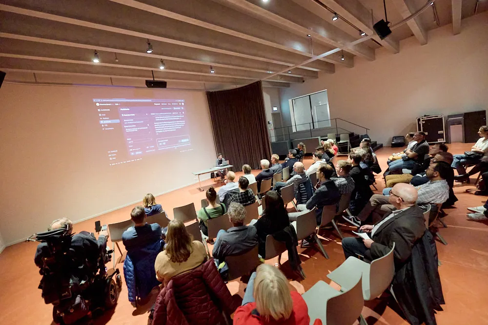 Ein Mann sitzt an einem Tisch vor einem Laptop. Er hält so eine Präsentation vor einem Publikum. Hinter dem Mann wird sein Bildschirm des Laptops gross an die Wand projiziert.