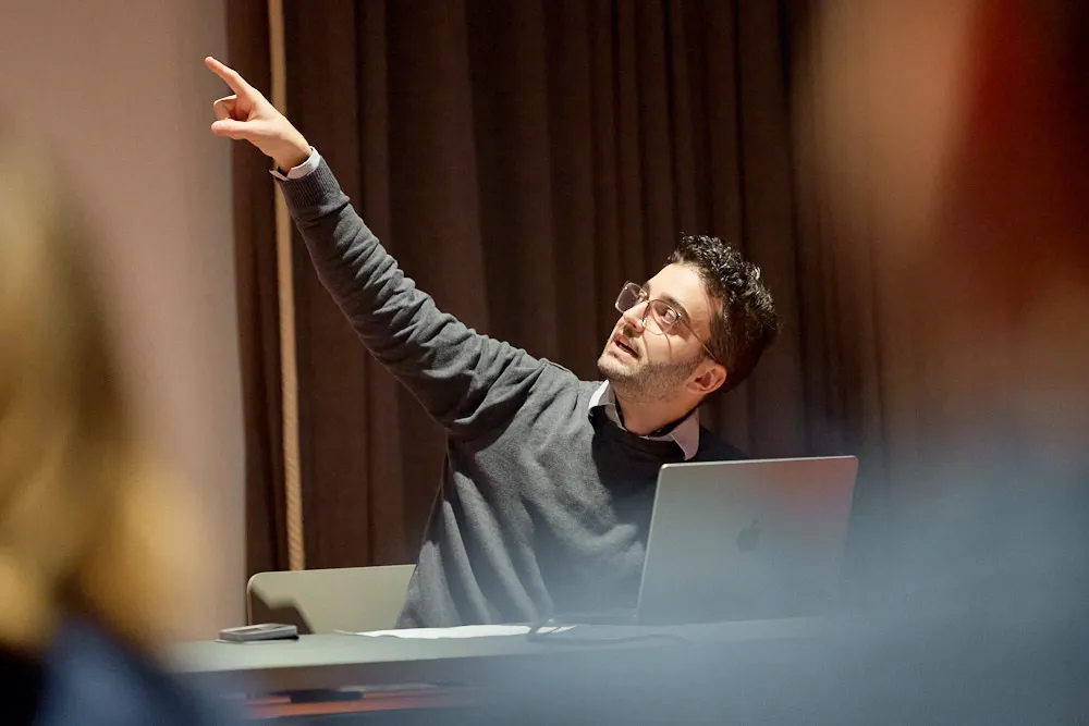 Ein Mann sitzt an einem Tisch vor einem Laptop. Er hält eine Präsentation. Er zeigt mit der rechten Hand auf die projizierte Präsentation an der Wand hinter ihm.