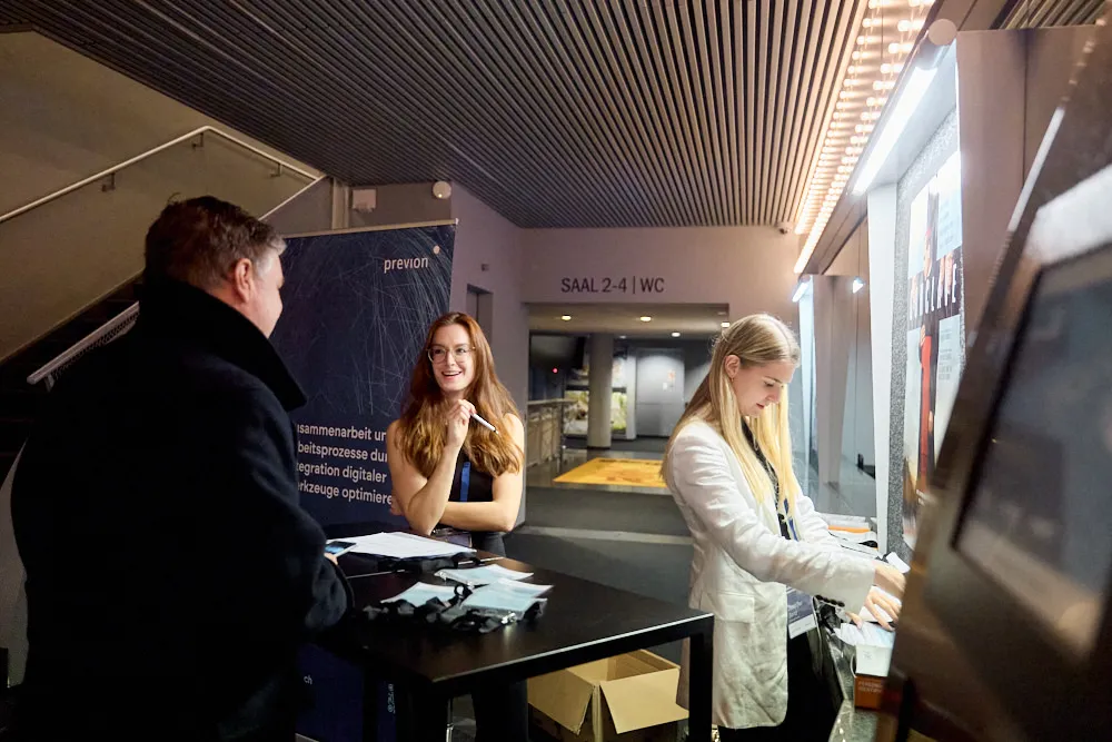 Zwei Frauen arbeiten am Einlass. Eine Frau lächelt dem männlichen Besucher vor ihr zu.