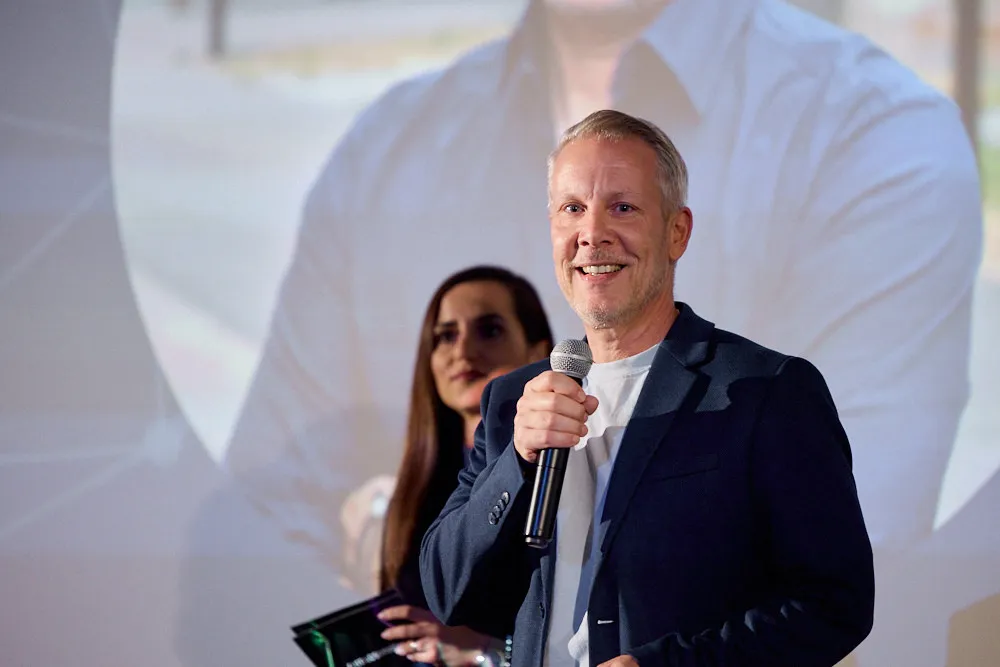 Ein Mann mit Mikrofon und eine Frau hinter ihm stehen vor einer Kinoleinwand.