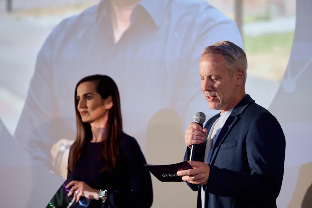 Ein Mann und eine Frau stehen vor einer Kinoleinwand. Sie halten beide Karten in der Hand. Der Mann hält zusätzlich ein Mikrofon in der Hand und spricht in es.