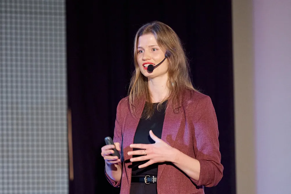 Eine Frau mit Headset spricht zu einem Publikum. Sie lächelt.