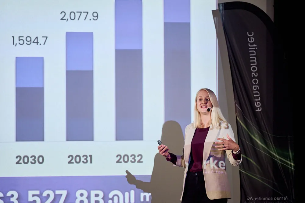 Eine Frau mit Headset steht vor einer Kinoleinwand und spricht. Auf der Leinwand ist eine Statistik ersichtlich.