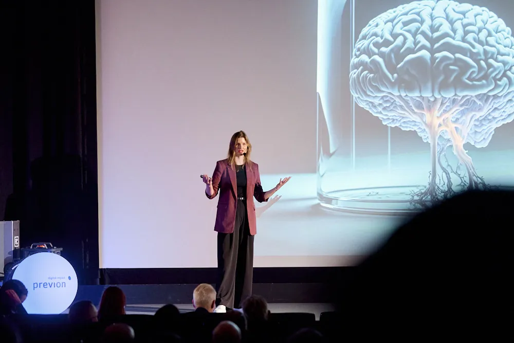 Eine Frau mit Headset steht vor einer Kinoleinwand und spricht. Auf der Leinwand ist ein Gehirn ersichtlich.