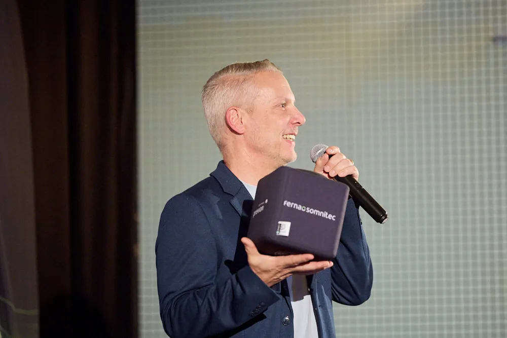 Ein Mann mit Mikrofon hält ein Stoffwürfel mit Logo darauf in der rechten Hand. In der linken Hand hält er ein Mikrofon. Er lächelt.