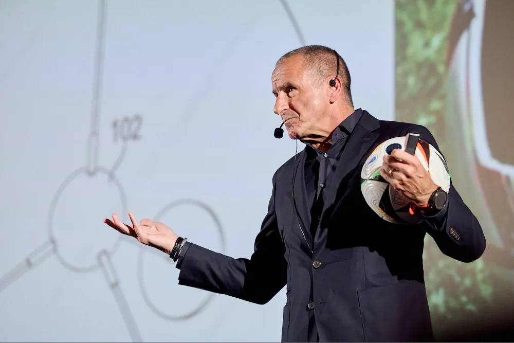 Ein Mann mit Headset hält eine Präsentation. In der einen Hand hält er einen Fussball.