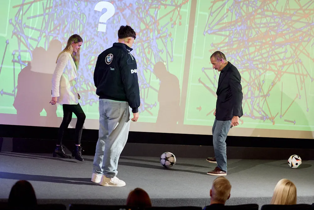 Zwei Männer und eine Frau spielen sich einen Fussball vor der Kinoleinwand zu.