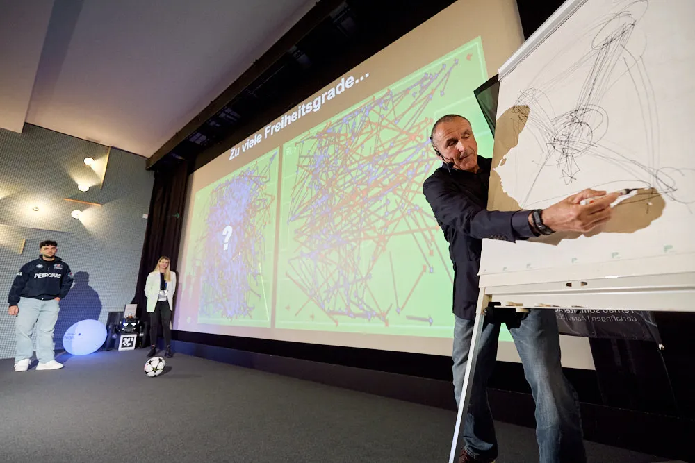 Ein Mann steht vor der Kinoleinwand und skizziert auf ein Whiteboard. Au der anderen Seite der Leinwand stehen ein Mann und eine Frau mit einem Fussball.