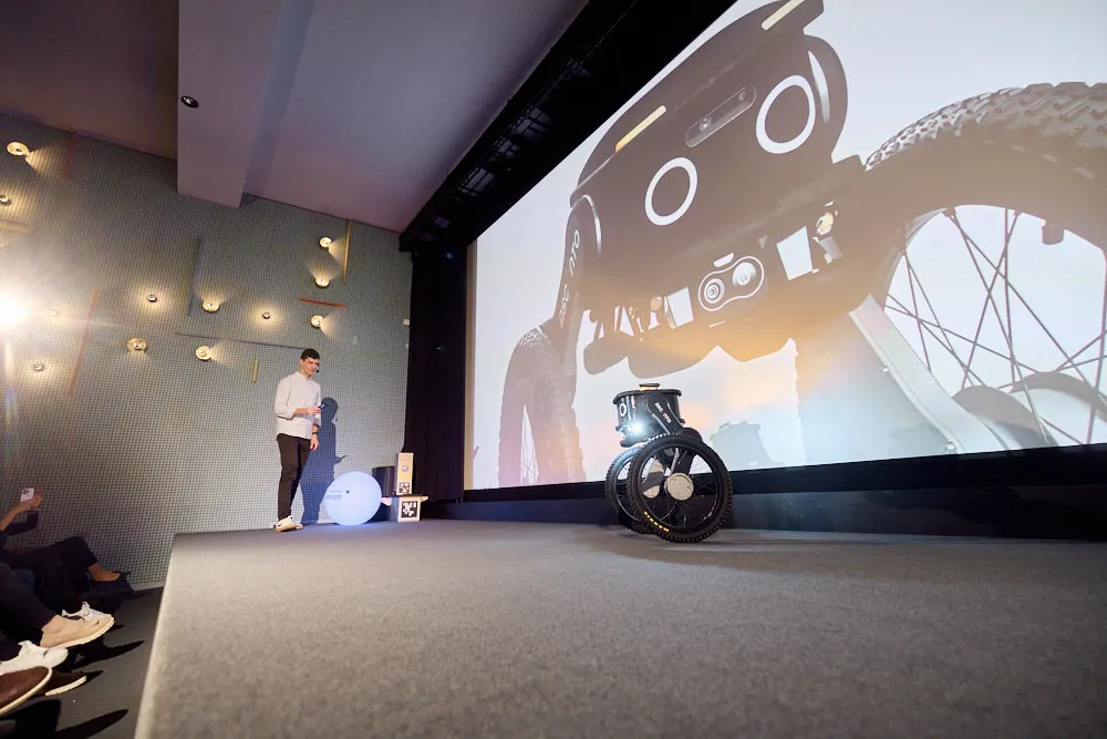 Ein Mann steht neben der Kinoleinwand und spricht zum Publikum im Kinosaal. Auf der Leinwand wird seine Präsentation projiziert. Vor der Leinwand befindet sich ein Roboter.