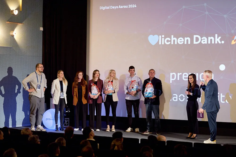 Es stehen fünf Frauen und vier Männer vor einer Kinoleinwand. Zwei Männer und zwei Frauen halten je ein Geschenk in den Händen.