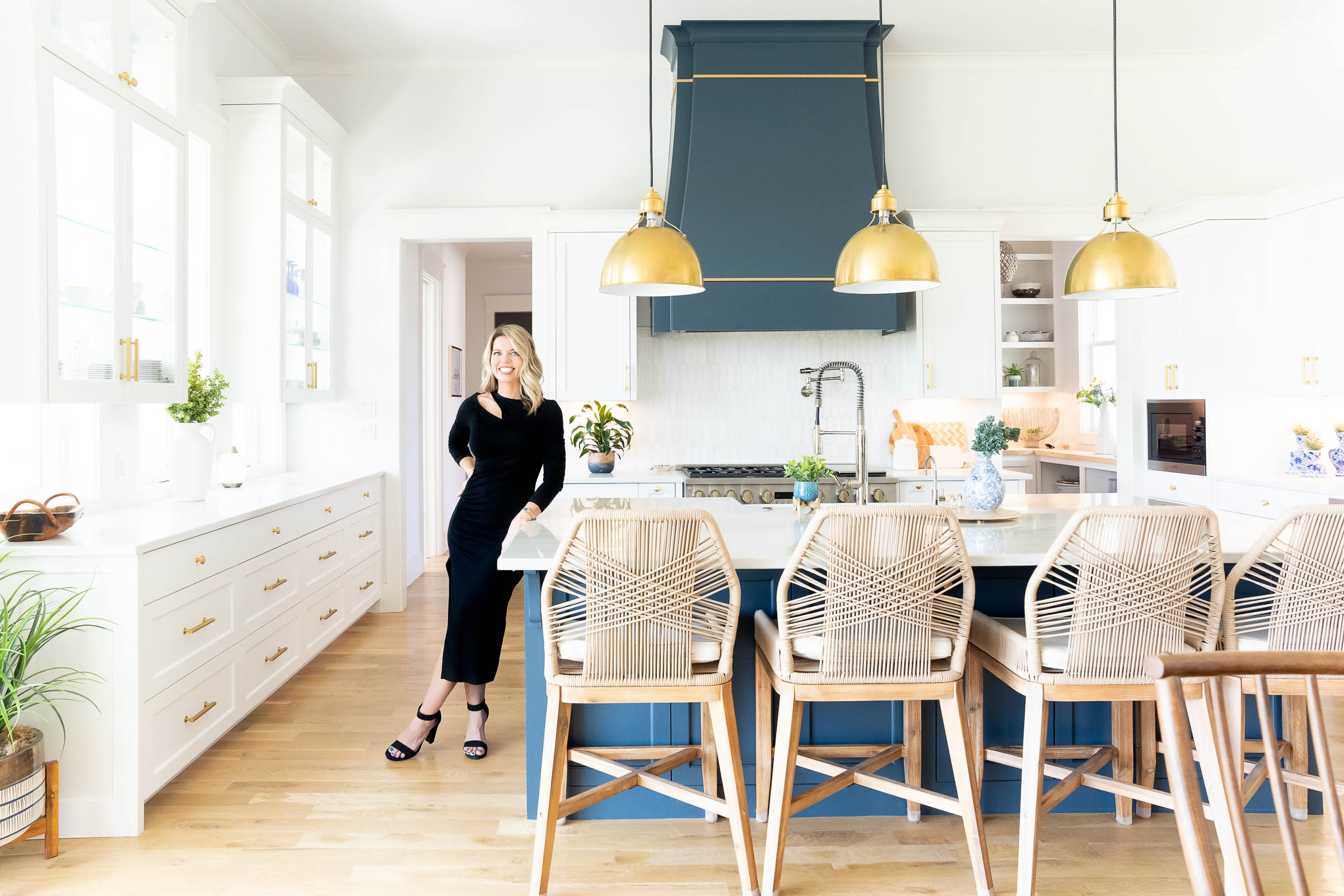 Woman in black dress standing and smiling in a bright white kitchen with blue island and gold pendant lights.