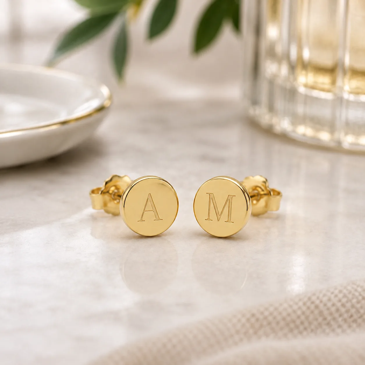 Pair of gold stud earrings engraved with the initials "A" and "M" on a marble surface