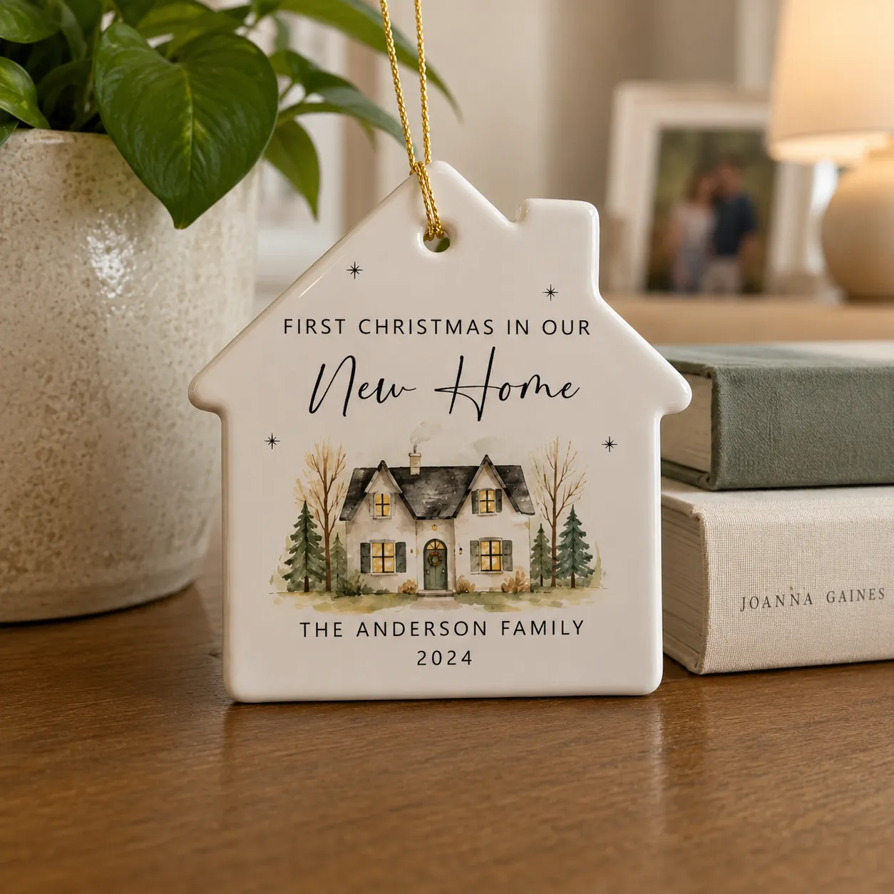 Ceramic house-shaped ornament reading "First Christmas in our New Home, The Anderson Family, 2024" on a table with books and a plant