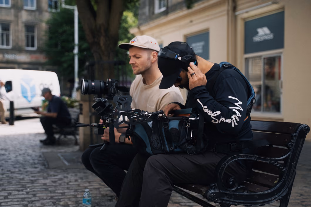 Two men sitting on a bench on a cobblestone street, one operating a camera rig and the other wearing headphones and handling equipment inside a bag.