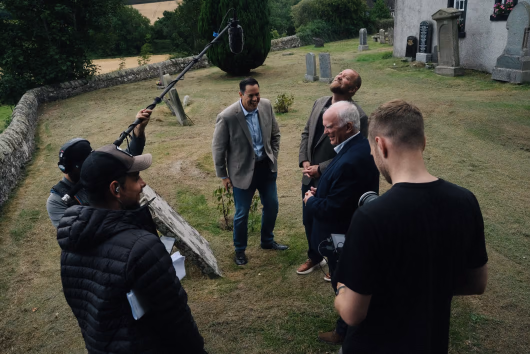 A small film crew records three men talking and laughing in a grassy graveyard with headstones, surrounded by trees and stone walls, one crew member holding a boom microphone overhead and another operating camera equipment.