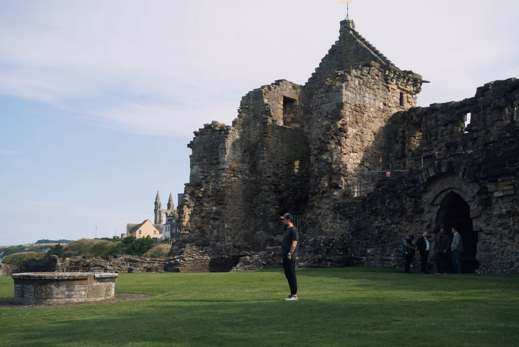 A man in casual clothing stands on green grass near the ruins of a large stone castle with an arched entrance, while four other people gather in the shadow of the castle wall under a partly cloudy sky.