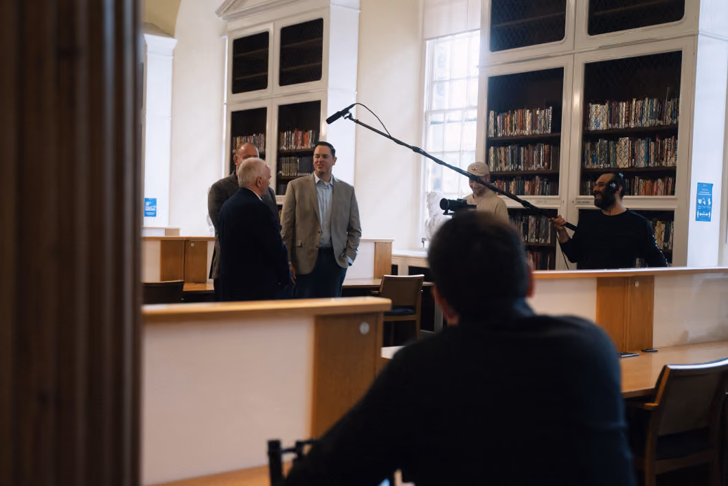 Three men standing and conversing near bookshelves in a well-lit room while a film crew records them; one person holds a camera and another operates a boom microphone, with a blurred person in the foreground facing them.