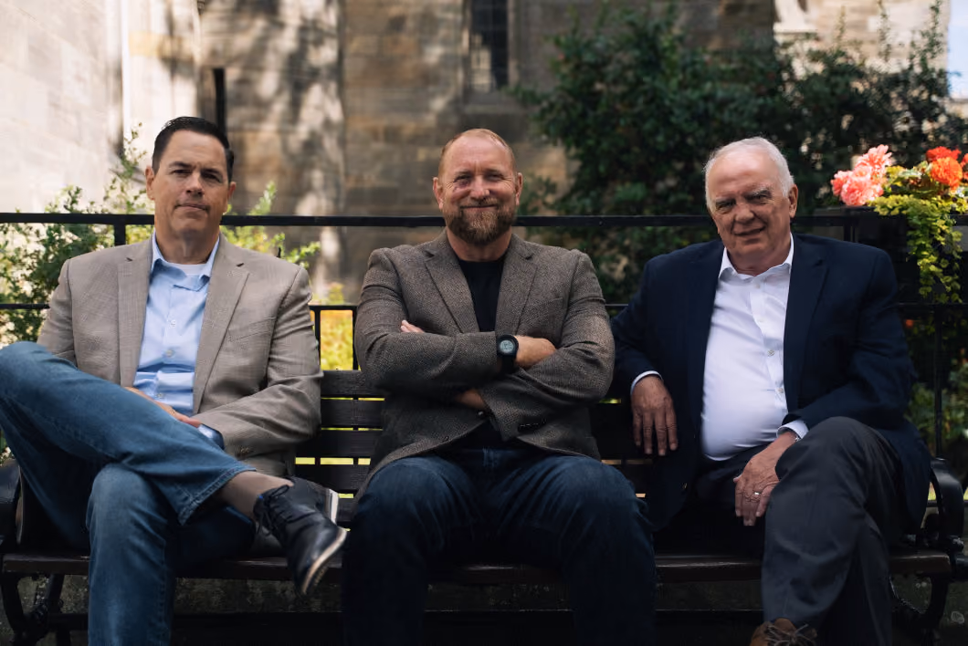 Three men sitting on a bench outdoors in front of stone architecture and greenery, wearing business casual attire with two men in blazers and one in a tweed jacket, one man in the center smiling with arms crossed.