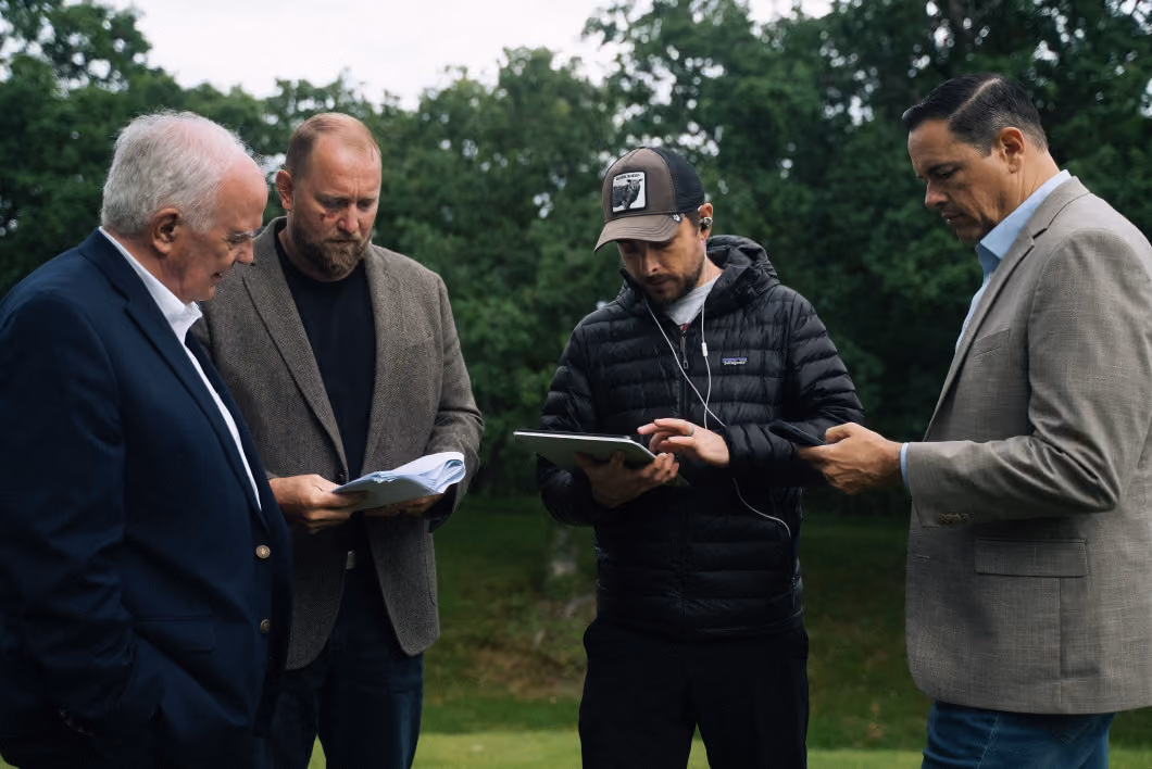 Four men outdoors in a green park area; one holding papers, another using a tablet with earphones, and another looking at a phone, all engaged in discussion or work.
