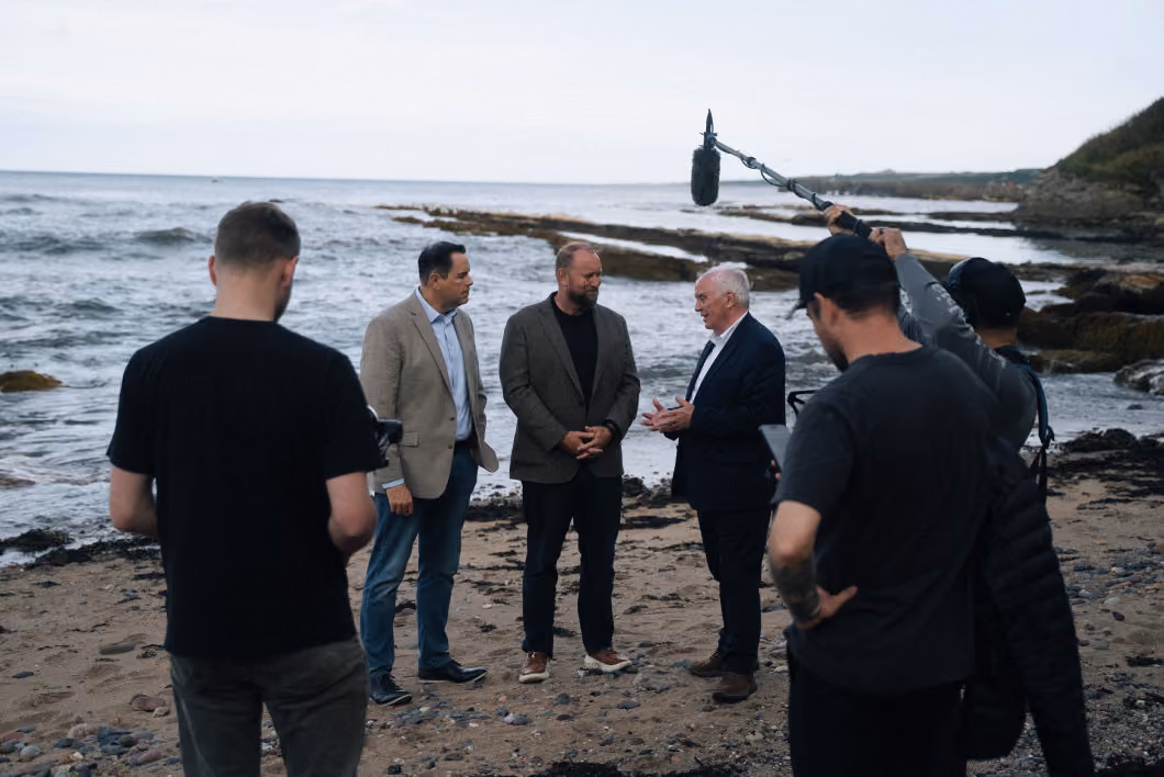 Three men dressed in business casual and formal attire stand on a rocky beach near the water, engaged in conversation while a film crew records them with a video camera and a boom microphone.