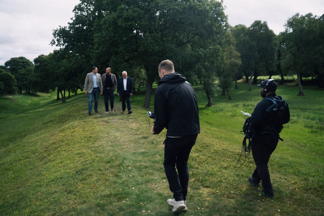 Two men walking on a grassy path among trees, conversing, while two crew members in black jackets and gear appear to film or record them.