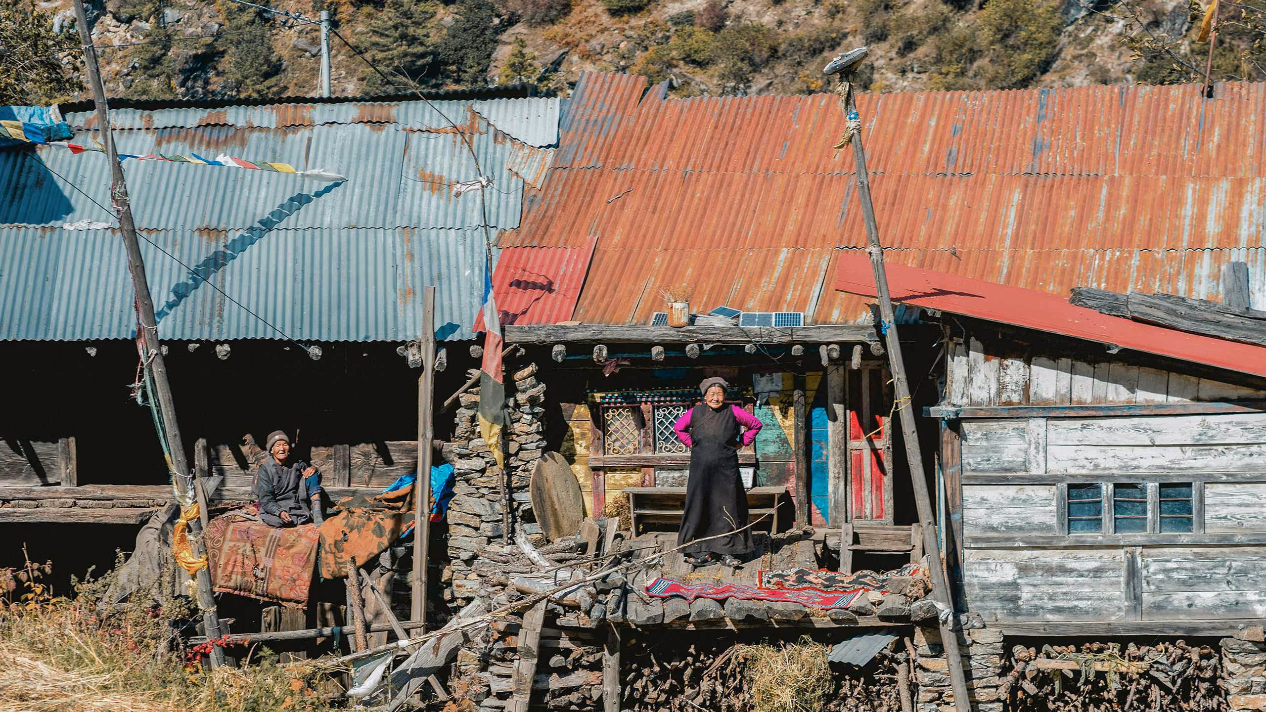 Two elderly people outside a rustic wooden and stone house with a corrugated metal roof; one person stands on a veranda with colorful walls, the other sits nearby on a makeshift seat, surrounded by dried plants and wooden poles.