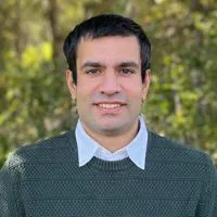 A person wearing a green sweater over a white collared shirt, standing outdoors with trees in the background.