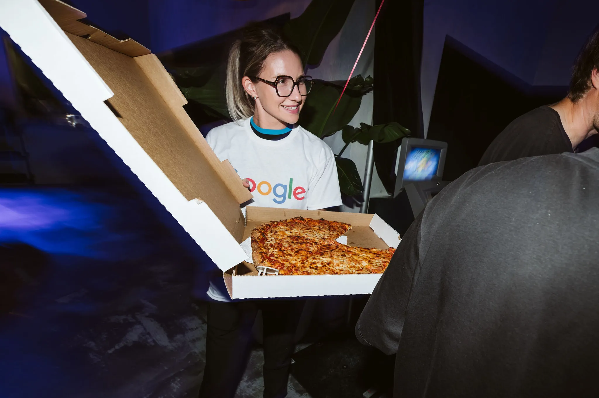 Une femme portant un t-shirt Google sourit en tenant une boîte ouverte contenant une pizza avec une tranche manquante.