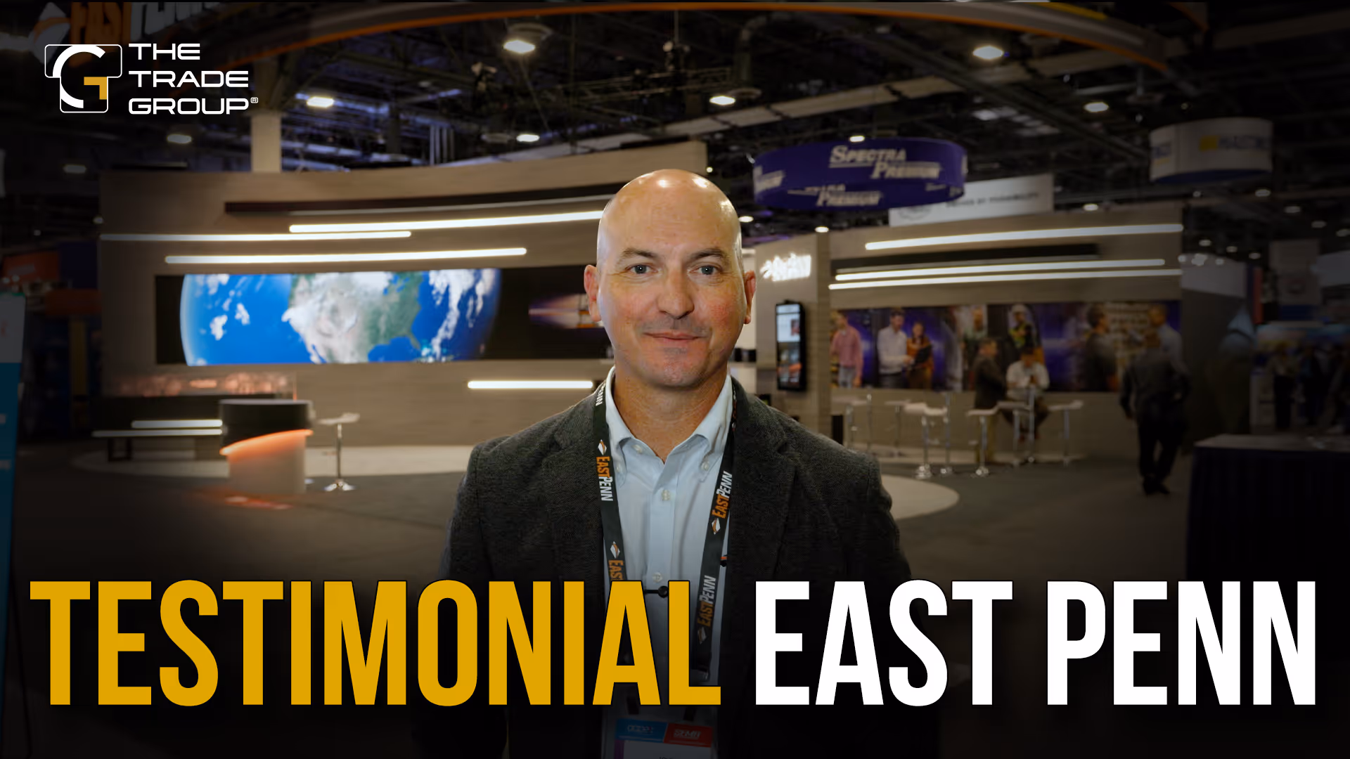 Man in business attire wearing an East Penn lanyard at a trade show booth with The Trade Group logo.