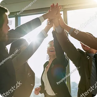 Business team in formal attire giving a group high five in an office with bright sunlight.