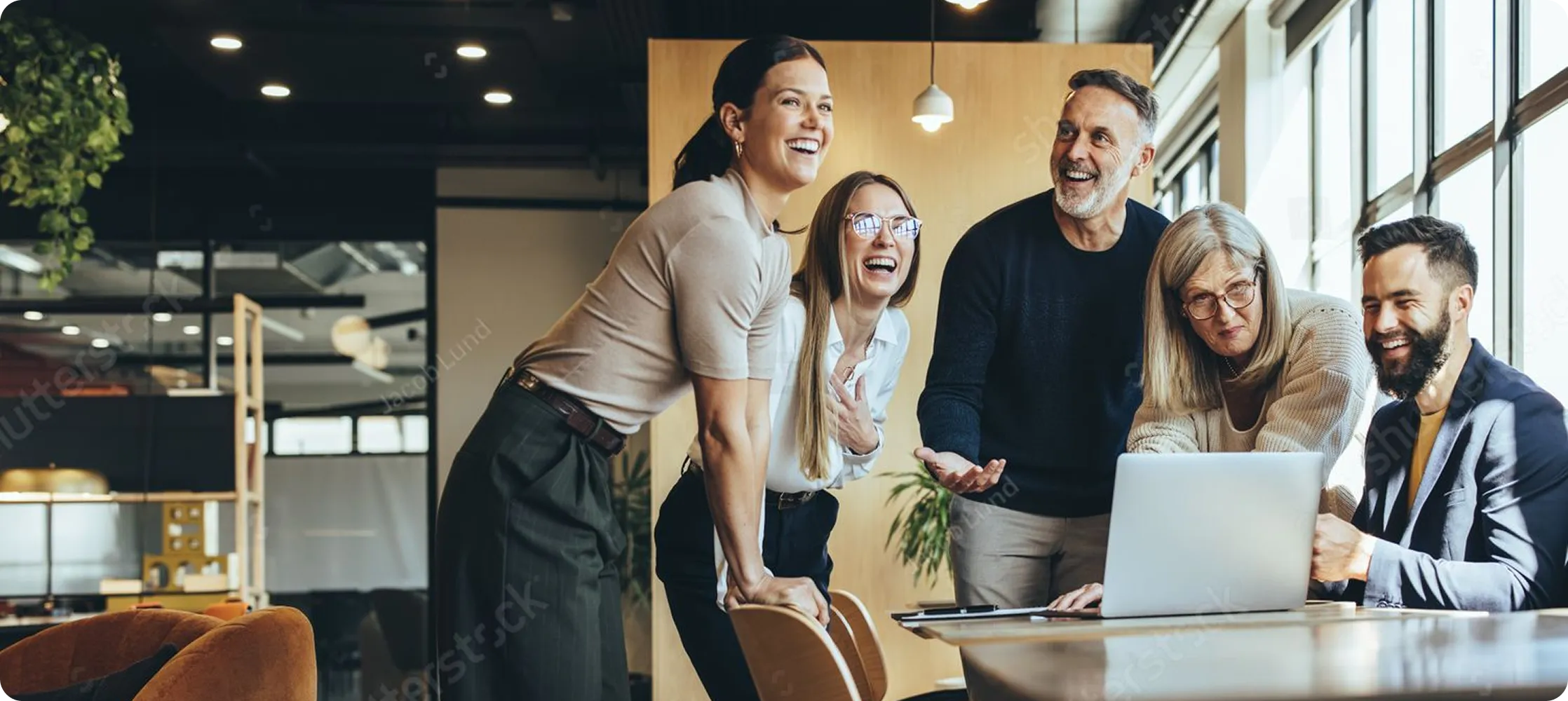 Five diverse professionals gathered around a laptop in a modern office, smiling and engaging collaboratively.