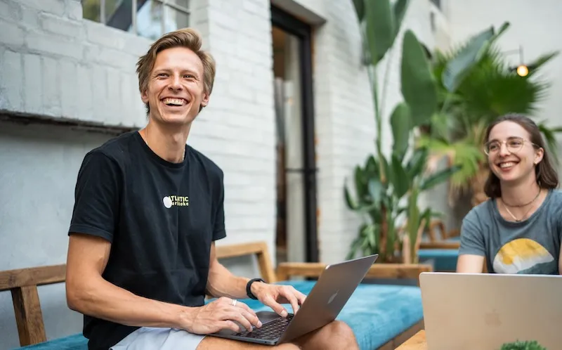 A smiling man works on a laptop outdoors beside a woman, both casually dressed and seated on a wooden bench near green plants.