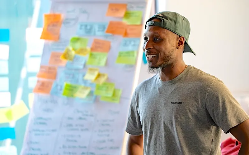 A man in a grey t-shirt and cap smiling during a brainstorming session in front of a flipchart filled with colorful sticky notes.