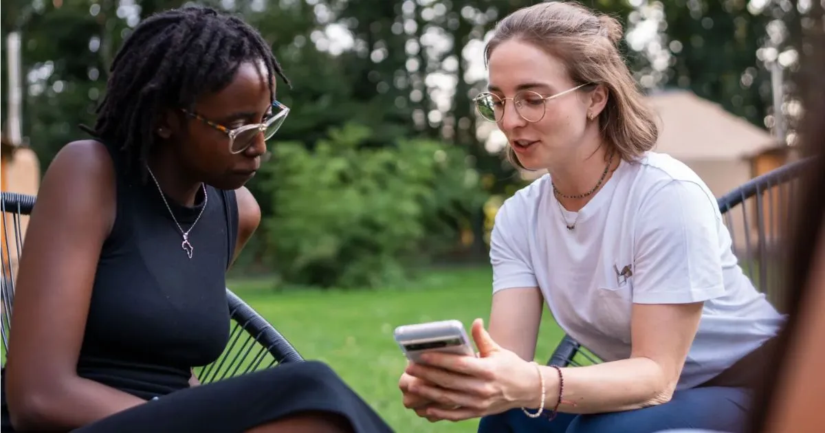 Two women are sitting outdoors on modern chairs, looking at a smartphone together. One is wearing a black dress with glasses and has braided hair; the other wears a white t-shirt and glasses, showing something on her phone. Trees and greenery are visible in the background.