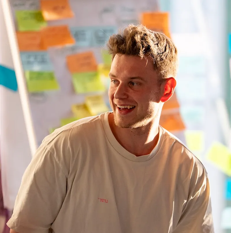 A young man laughing while presenting with a kanban board in the background.