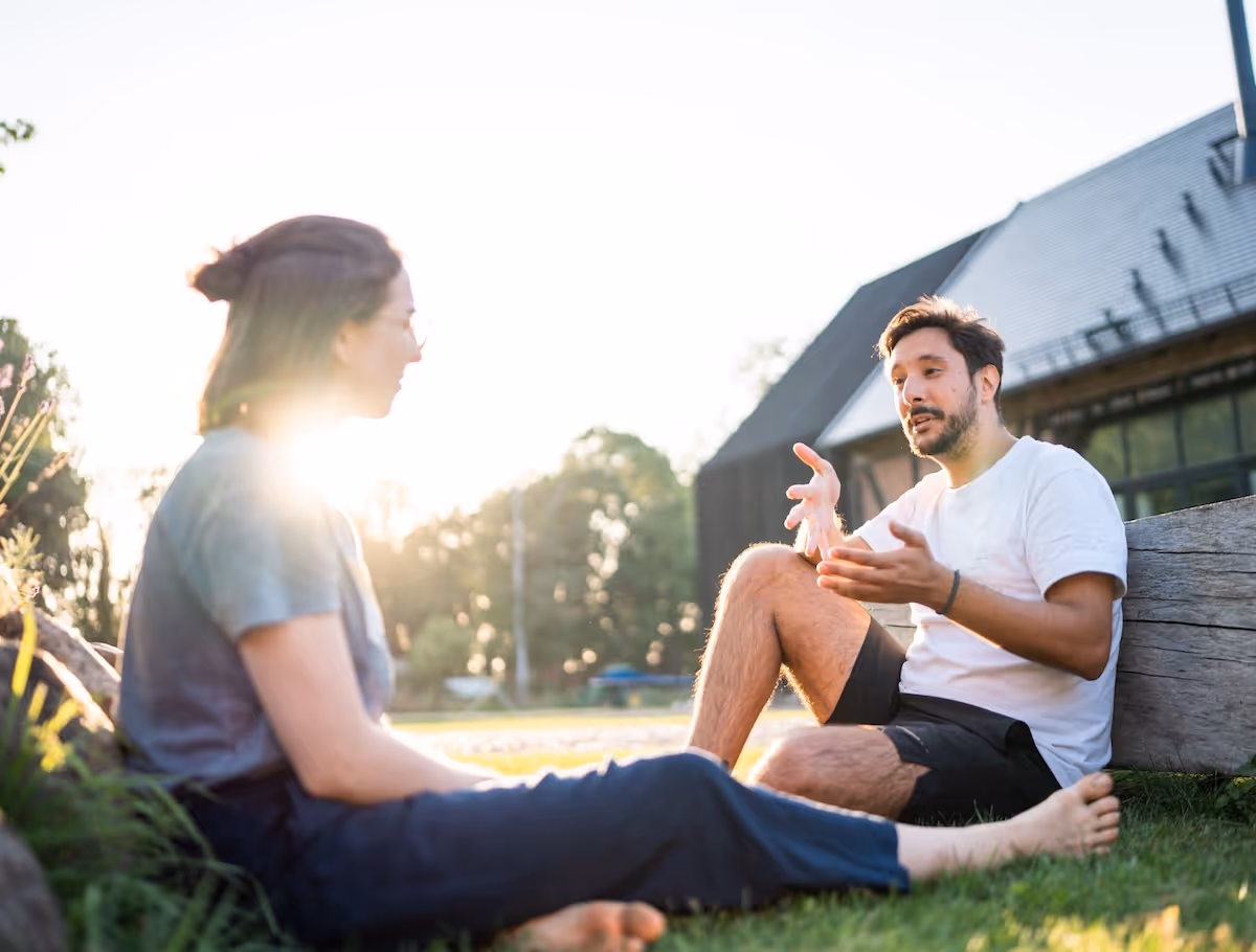 Two people sitting on grass outdoors, having a conversation with sunlight behind them.