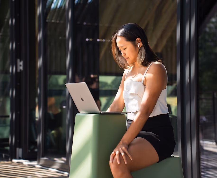 Woman sitting outdoors working on a laptop placed on a green cube-shaped stand, wearing wireless earbuds.