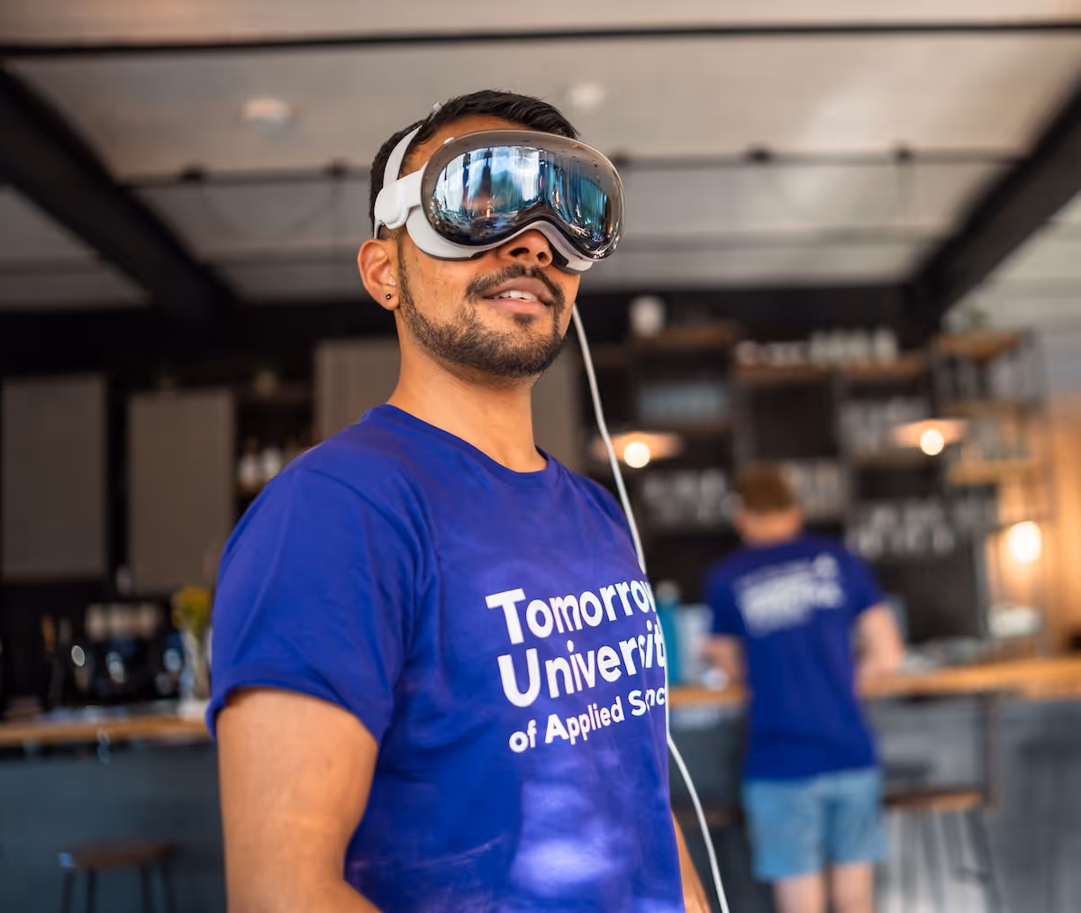 Man wearing a blue 'Tomorrow University of Applied Sciences' T-shirt and virtual reality goggles in an indoor setting with another person in the background.