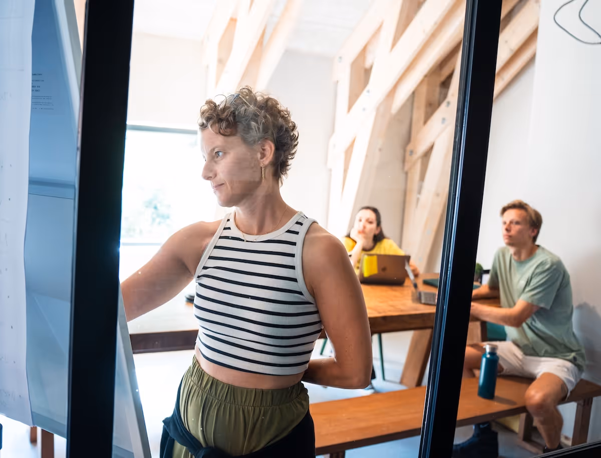 Woman with short curly hair writing on a whiteboard while two colleagues watch attentively in a modern office room.