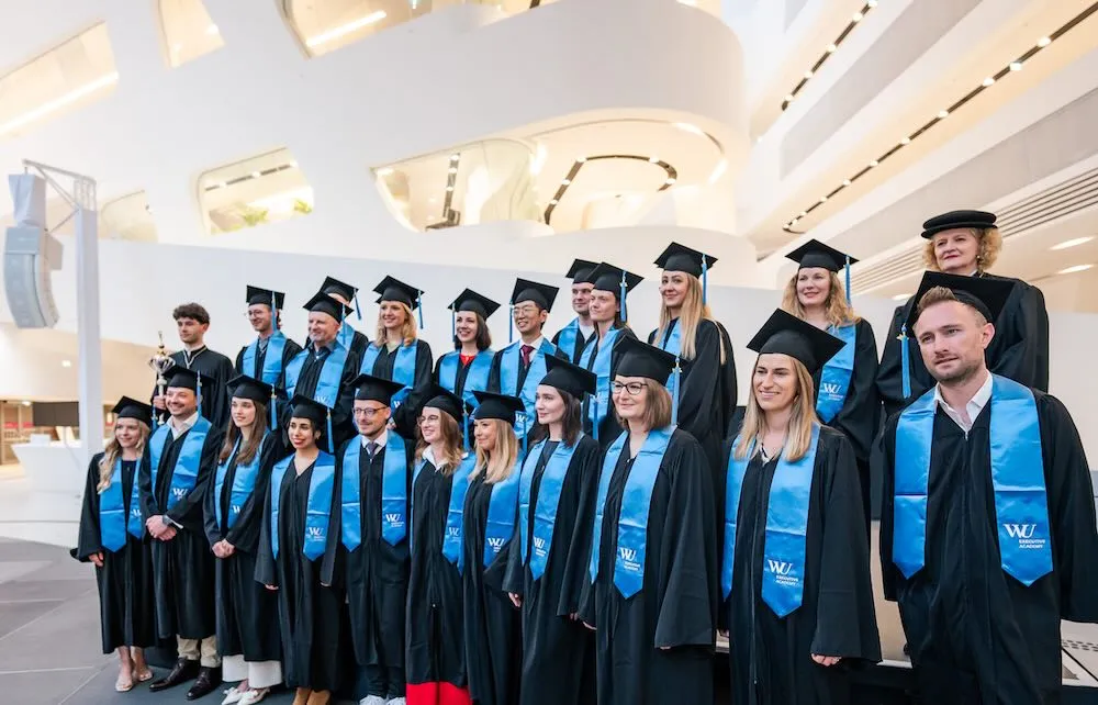 Group of diverse graduates wearing black caps and gowns with blue stoles posing indoors in a modern building.