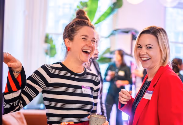 Zwei lächelnde und lachende Frauen bei einem geselligen Beisammensein drinnen, eine trägt ein gestreiftes Oberteil und die andere einen roten Blazer.