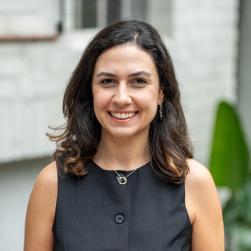 Smiling woman with shoulder-length dark hair wearing a sleeveless black top and a circular pendant necklace, standing outdoors.