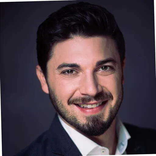 Smiling man with dark hair, a short beard, and mustache wearing a dark suit jacket and white shirt.