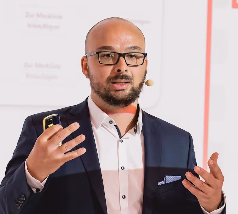 Man with glasses and beard speaking, wearing a suit, white shirt, and headset microphone.
